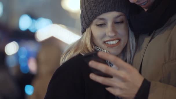 Pareja joven viendo fotos en el teléfono inteligente por la noche en una calle de la ciudad . — Vídeos de Stock