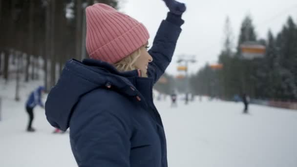 Młoda kobieta uśmiechający się w odzieży zimowej, czekając na jej przyjaciół, w pobliżu wyciągu narciarskiego. — Wideo stockowe