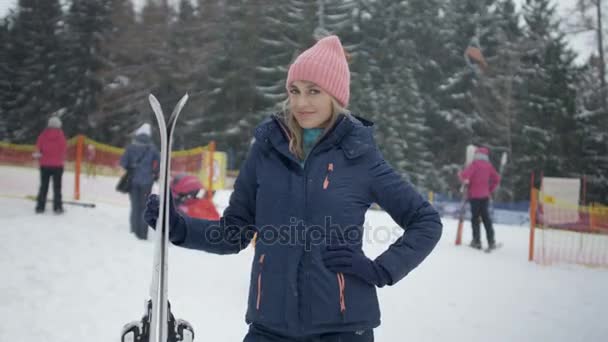 Ung glad kvinna i vinterjacka som håller ski mot skidområde. — Stockvideo
