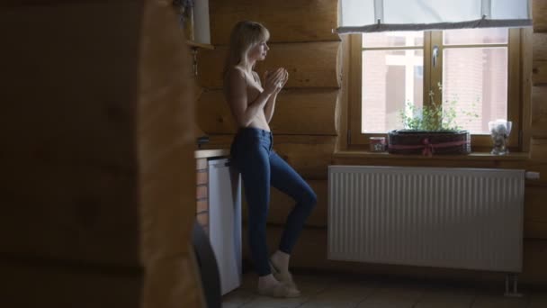 Schöne junge nachdenkliche Frau hält eine Tasse heißen Tee in der Hand und steht am Fenster. — Stockvideo