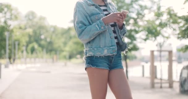 Mooie jonge vrouw die het dragen van denim jasje te typen op de telefoon in een stadspark tijdens zonnige dag. — Stockvideo