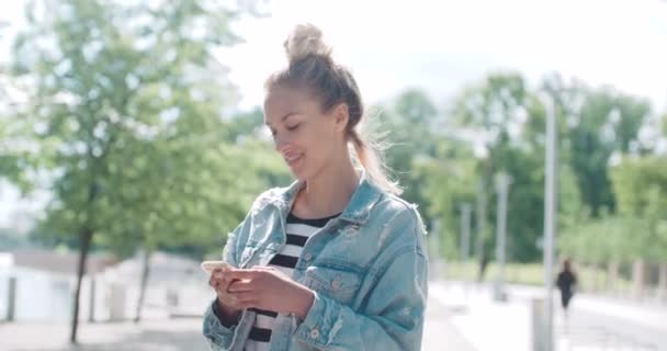 Hermosa mujer joven con chaqueta de mezclilla escribiendo en el teléfono en un parque de la ciudad durante el día soleado . — Vídeo de stock
