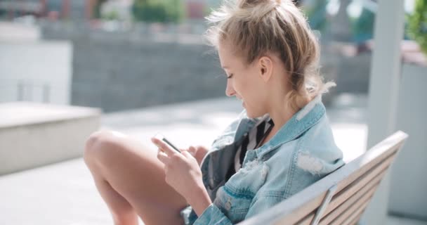 年轻漂亮的女孩坐在木凳上，使用手机上. — 图库视频影像