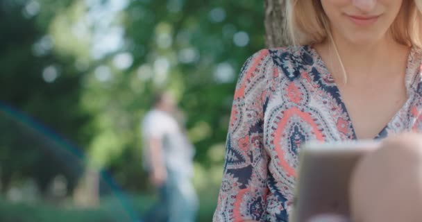 Молодая женщина сидит в парке и использует цифровой планшет . — стоковое видео
