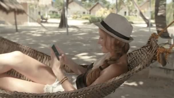 在热带度假胜地使用电话的迷人的妇女 美丽的女人坐在吊床上 在电话上打字 — 图库视频影像