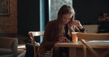 Restoranda oturan ve portakal suyu içme sırasında cep telefonu kısa mesaj yazarak neşeli bir genç kadının portresi.