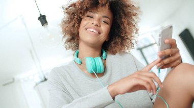 Müzik keyfi ve telefon tutarak mutlu ve neşeli genç kadın. Güzel genç afro Amerikalı kadın evde mutfak masasında oturan renkli kulaklıklarla.