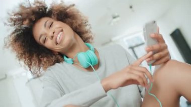 Müzik keyfi ve telefon tutarak mutlu ve neşeli genç kadın. Güzel genç afro Amerikalı kadın evde mutfak masasında oturan renkli kulaklıklarla.