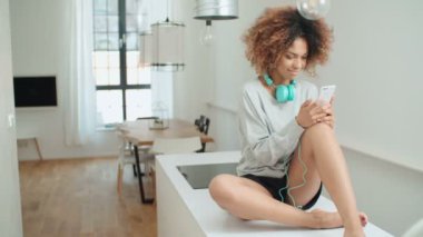 Müzik keyfi ve telefon tutarak mutlu ve neşeli genç kadın. Güzel genç afro Amerikalı kadın evde mutfak masasında oturan renkli kulaklıklarla.