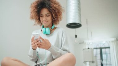 Müzik keyfi ve telefon tutarak mutlu ve neşeli genç kadın. Güzel genç afro Amerikalı kadın evde mutfak masasında oturan renkli kulaklıklarla.