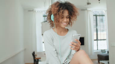 Müzik keyfi ve telefon tutarak mutlu ve neşeli genç kadın. Güzel genç afro Amerikalı kadın evde mutfak masasında oturan renkli kulaklıklarla.