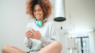 Müzik keyfi ve telefon tutarak mutlu ve neşeli genç kadın. Güzel genç afro Amerikalı kadın evde mutfak masasında oturan renkli kulaklıklarla.