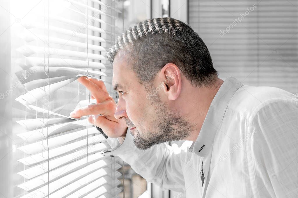 Man in isolation looking through window blinds. Coronavirus outbreak. Man looks at a sunny street through the window blinds