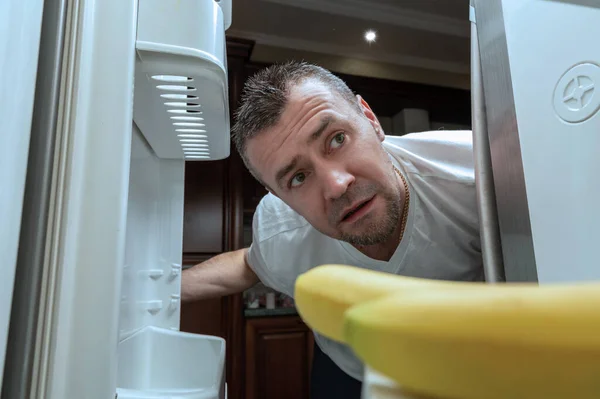 Man peers into an empty refrigerator during self-isolation. Shocked man looking into empty refrigerator at home. COVID-19. Coronavirus consequence