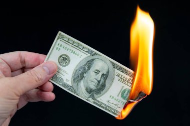 100 dolarlık banknotu yakmak. Yanan banknotu tutan bir el. Yaklaşık dörtte biri büyük bir alev tarafından tüketiliyor. Siyah arkaplan.