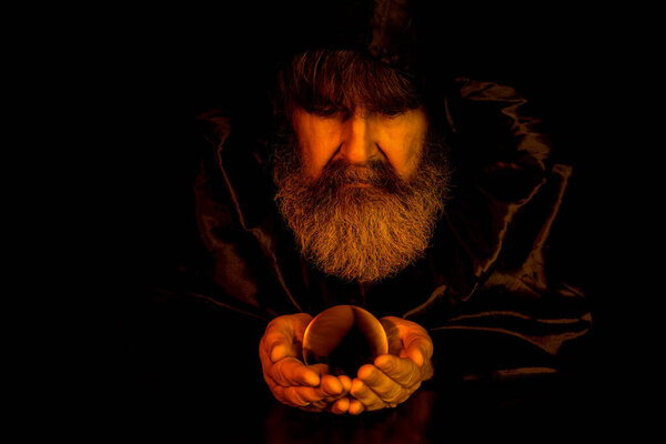Wizard holding a crystal ball in his hands. He wears a dark cloak. Shallow depth of field, focus on face. Dark scene illuminated by light from fire.
