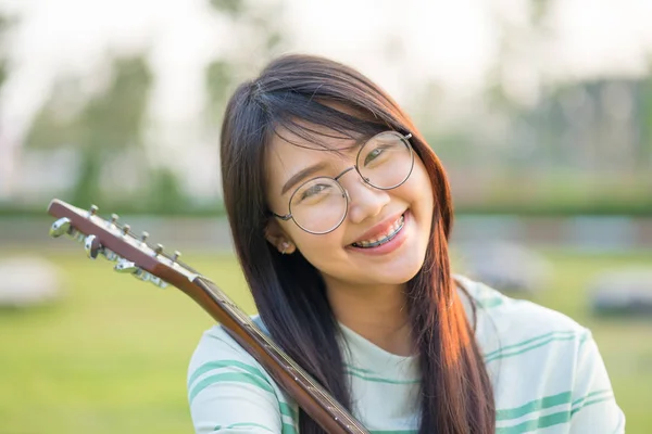 在草坪上肩膀上有吉他的亚洲少女 她戴着大括号戴着眼镜 — 图库照片