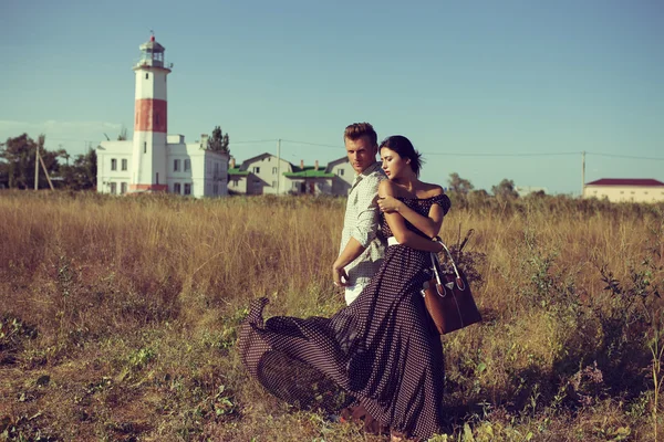 young couple hipster indie style in love walking in countryside, holding hands, lighthouse on background, warm summer day, sunny, bohemian outfit, vintage bag with flowers