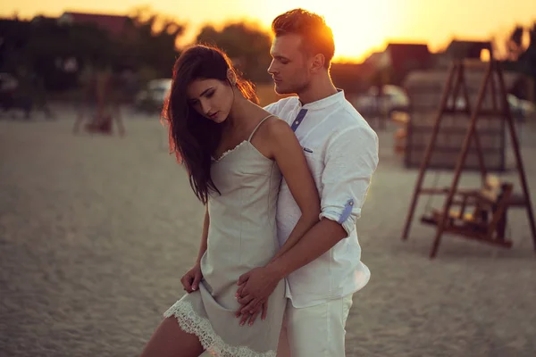Μοντέρνα νεαρό ζευγάρι που στέκεται, στηρίζεται πάνω στην παραλία, φορώντας ένα λευκό ρούχα θέτοντας αγάπη στο ηλιοβασίλεμα. με προσαρμοσμένη ισορροπία λευκού, φίλτρα χρώματος, και μερικά ωραία ταινία θόρυβος προστίθεται στην εικόνα — Φωτογραφία Αρχείου
