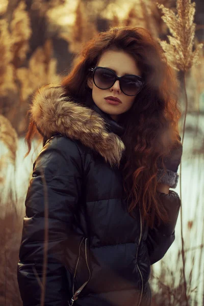 Fashion street stil utseende casual kläder för kvinnan promenader på park, varma textur svart jacka med päls huva vackra sexiga modell, vackert ansikte, rödhårig modell lyx livsstil vintern sunset bakgrund — Stockfoto