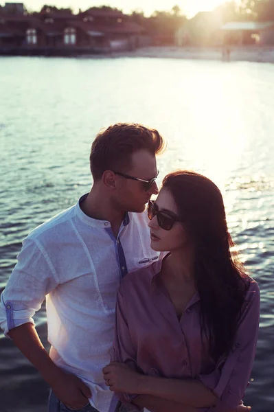 Sunset bulvarında deniz kenarında oturan genç mutlu şık çift. Adam ve kadın romantik seksi yaz giysileri ve şık moda güneş gözlüğü takmış — Stok fotoğraf