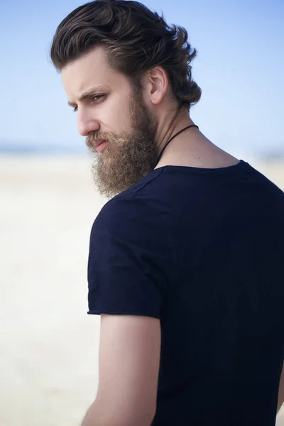 Retrato de um homem jovem e bonito com uma barba em t-short preto posando na costa do mar. Fecha. Conceito de moda mar / praia . — Fotografia de Stock