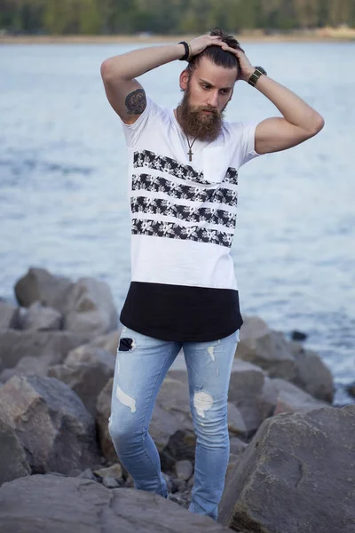 Retrato de um homem jovem e bonito com uma barba na moda t-short posando na costa do mar. Fecha. Conceito de moda mar / praia . — Fotografia de Stock