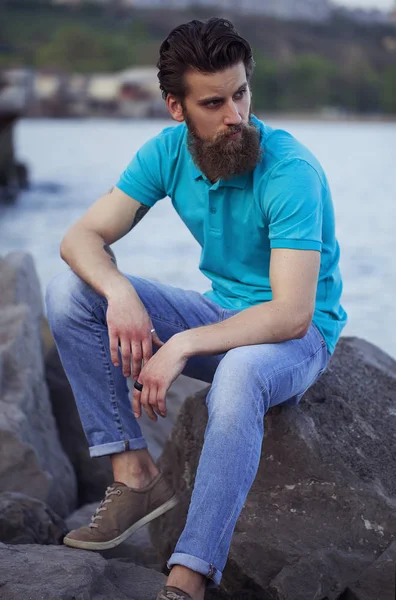Portréja egy fiatal és jóképű férfi szakáll, divatos t-rövid pózol a tenger partján. Közelről. Tengeri/tengerparti divat fogalma. — Stock Fotó