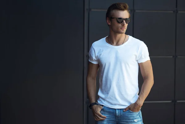 Street fashion concept. Portrait of a young and handsome man in a white t shirt and trendy glasses posing over black modern background. Close up. Street shot. copy space — Stock Photo, Image