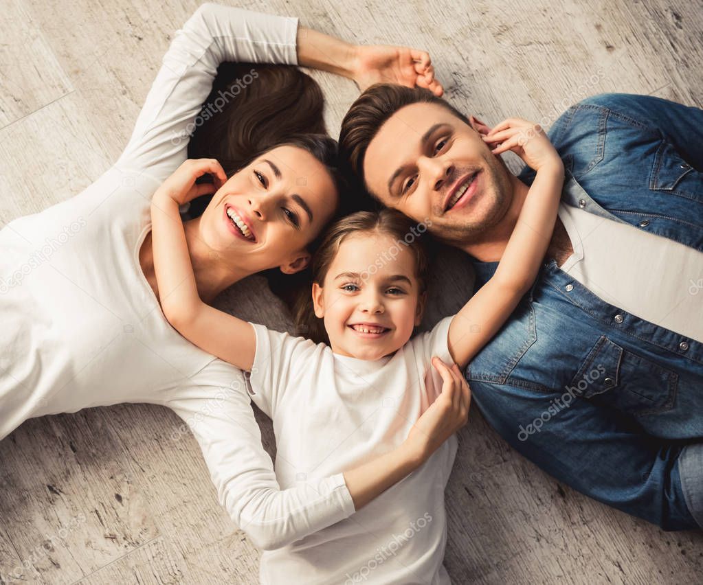Top view of cute little girl and her beautiful young parents looking at camera and smiling while lying on the floor at home