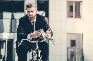 Yakışıklı sakallı işadamı Klasik takım elbiseli bir akıllı telefon kullanarak ve şehirde bisiklet sürme sırasında kameraya bakıyor