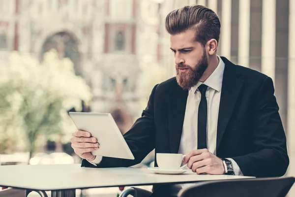 クラシックなスーツのハンサムなひげを生やしたビジネスマンはデジタル タブレットを使用して 市内中心部のカフェに座りながらコーヒーを飲んで — ストック写真