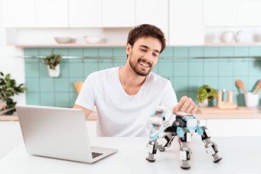 Bir adam mutfakta bir robot programlama. O gri bir dizüstü bilgisayarda çalışıyor. Robot tablo yanında duruyor. Robot küçük mekanik gergedan gibi görünüyor.