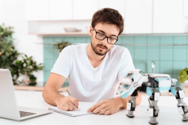 Bir adam mutfakta bir robot programlama. Yakınlarda robot kendisi ve gri bir dizüstü bilgisayar var. Bir adam bir şey bir not defterinde yazıyor.