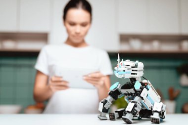 Kız mutfakta ayakta ve bir tablet tutarak. Bir küçük gergedan robot tablo yanında oturur. Bir kız ekrana bakarak ve gülümseyen.