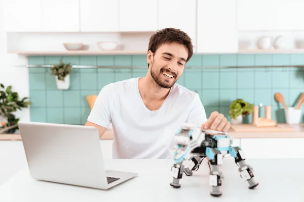 男は台所でロボットをプログラミングです 彼は灰色のノート パソコンで動作します ロボットは テーブルの横に立っています ロボットが小さな機械のサイのように見える — ストック写真