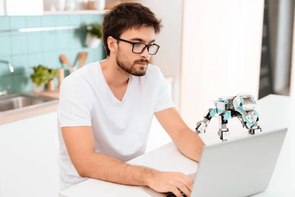 A man is programming a robot in the kitchen. He works on a gray laptop. The robot stands next to the table. The robot looks like a small mechanical rhinoceros.