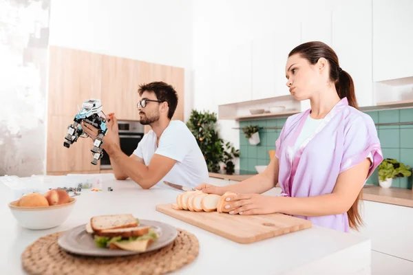 A woman is preparing breakfast. A man sits next to a table and collects a robot. He does not pay attention to the woman. She is indignant.