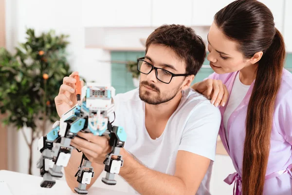 A man collects a robot in the kitchen. The woman affectionately speaks to him. The man shrugs it off. They are located in a light modern kitchen. He wears a home-made dressing gown on his wifes.