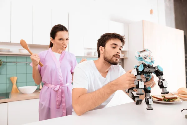 A man collects a robot in the kitchen. His girlfriend is behind him and very angry. She is dressed in a pink robe and yells at the guy.