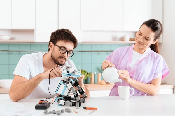 A man creates a robot in the kitchen. His girlfriend is standing by and pouring tea. They both smile. The girl watches how the guy works.