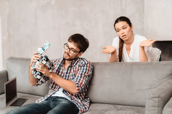 Young couple in the living room. The guy is holding a robot. The girl next to him, and she is very unhappy that the guy ignores her. They are sitting on the gray couch in the living room.