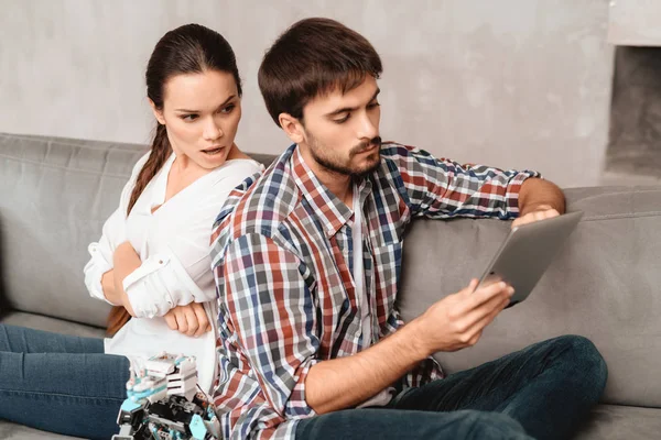 Young couple in the living room. The guy is holding a robot. The girl next to him, and she is very unhappy that the guy ignores her. They are sitting on the gray couch in the living room.