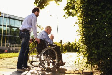 Yaşlı adam parkta tekerlekli sandalyede oturuyor. Onun arkasında onun oğlu duruyor. Onlar yürüyüş. Yaşlı adam üzücü