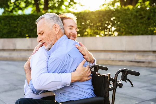 Ao velho que se senta no parque em uma cadeira de rodas veio seu filho. Ele está abraçando o velho. — Fotografia de Stock