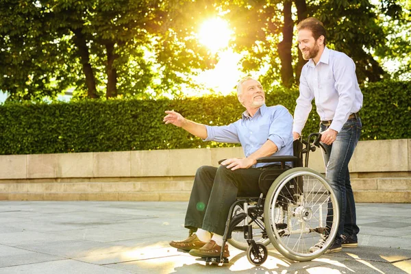 Un uomo cammina nel parco con suo padre, seduto su una sedia a rotelle. L'uomo e il vecchio sono felici — Foto Stock