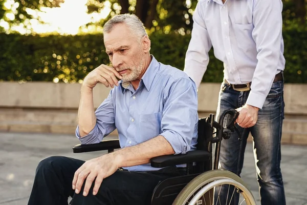 Een man loopt in het park met zijn vader, die in een rolstoel zit. De oude man is triest — Stockfoto