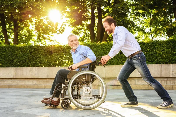 Un uomo sta cavalcando suo padre su una sedia a rotelle nel parco. Sono felici. — Foto Stock