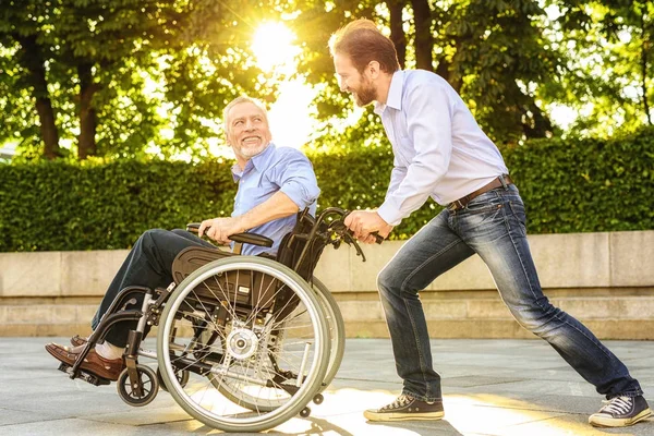 Un hombre está montando a su padre en una silla de ruedas en el parque. Son felices. —  Fotos de Stock