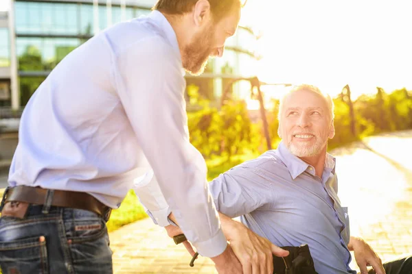 Der alte Mann sitzt im Rollstuhl im Park. Hinter ihm steht sein Sohn. der alte Mann schaut glücklich seinen Sohn an — Stockfoto